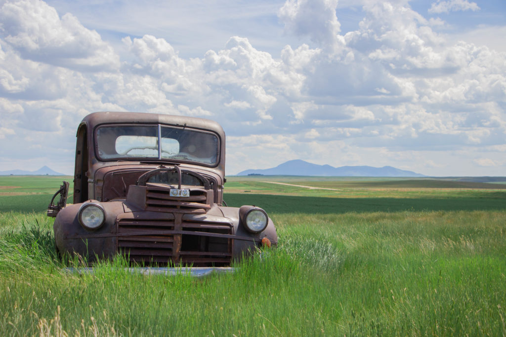 Rusty old truck in tall green grass with blue sky and clouds and hills mountains in background