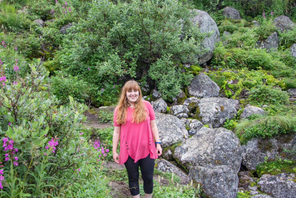 Girl in pink shirt and leggings standing by rocks, green plants and flowers, and mountain stream in Hatcher Pass Alaska